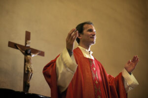 un prêtre en chasuble rouge prêche les bras écartés, dos à une croix, dans un bâtiment.