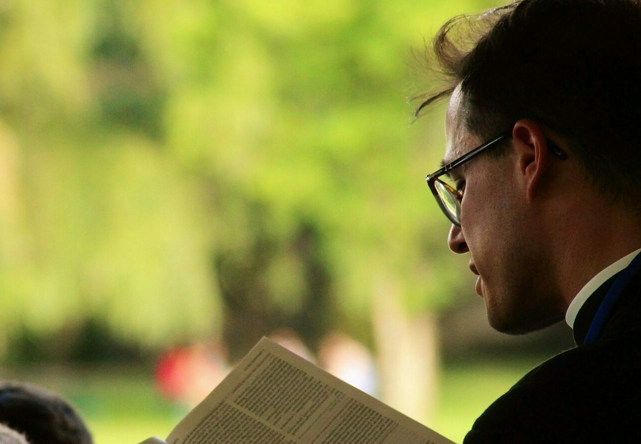 un prêtre catholique debout au premier plan, lit une lecture dans la Bible à une assemblée de dos, assise. La scène semble se dérouler sous un couvert, à l'extérieur. Il fait beau.